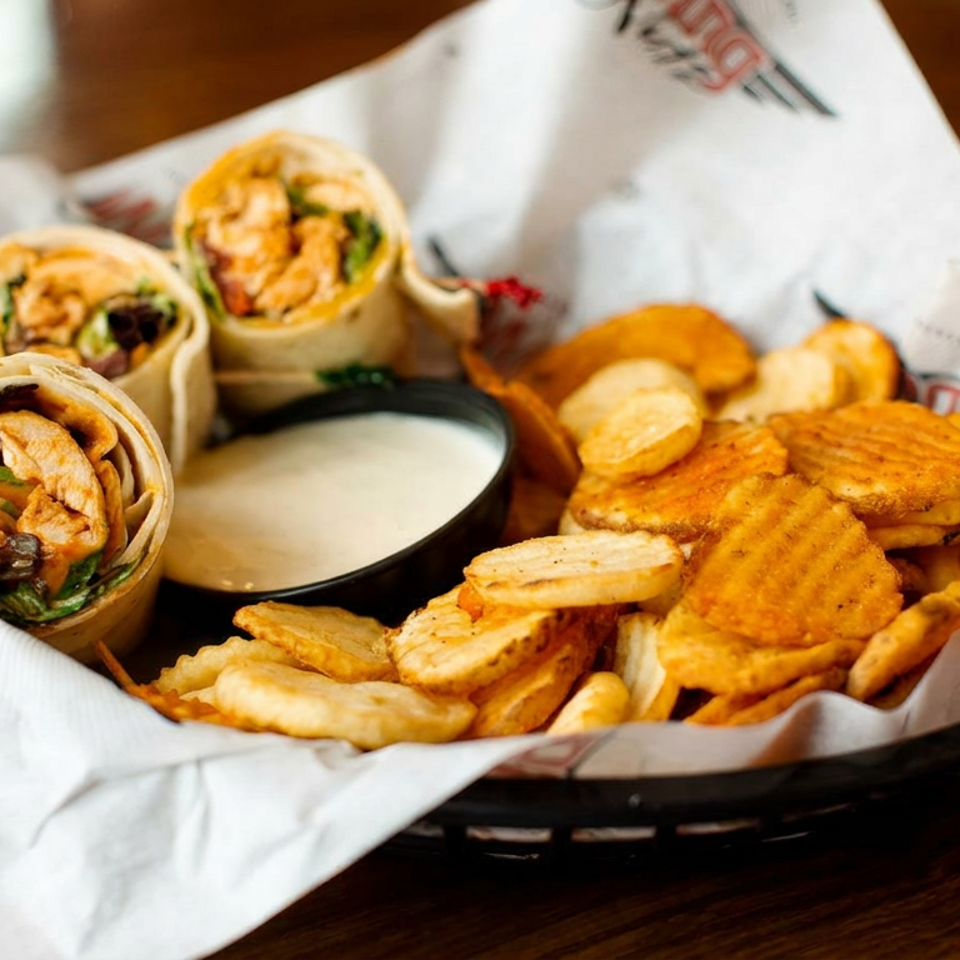 Basket of grilled chicken wraps, crispy chips, and dipping sauce served on a wooden restaurant table.