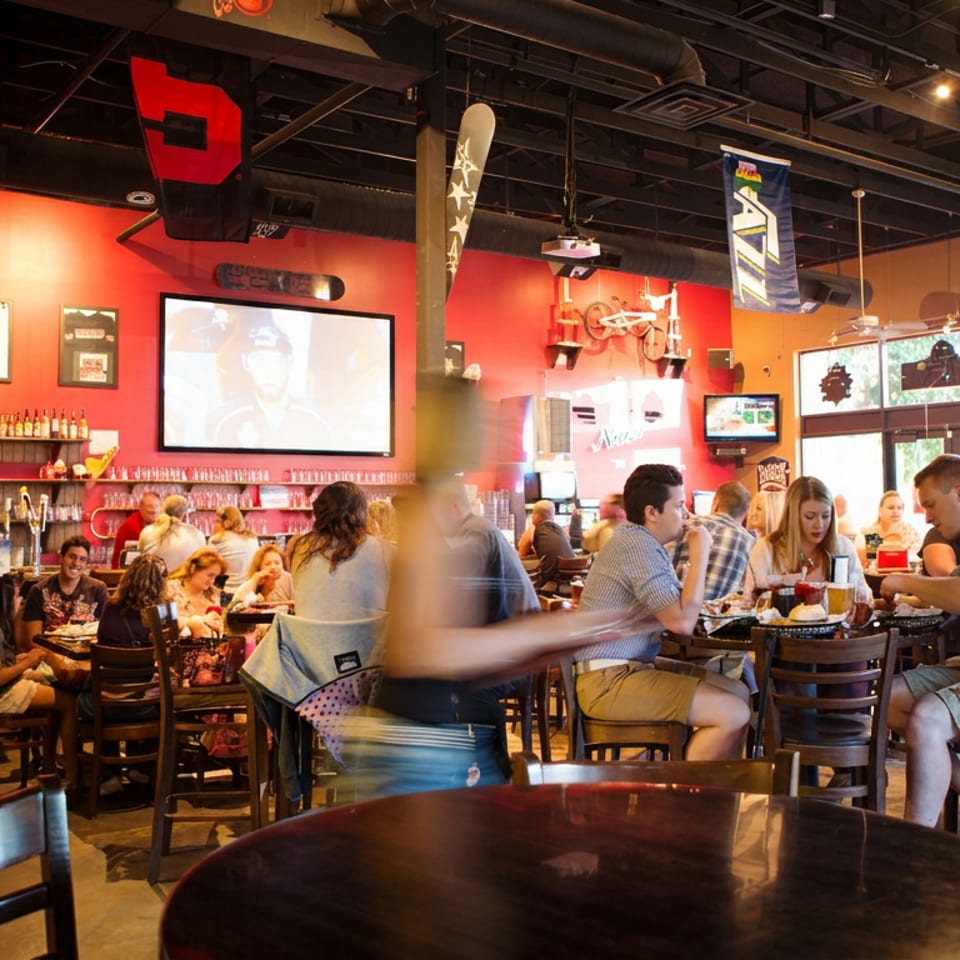 Bustling sports bar restaurant with red walls, televisions, hanging banners, a stocked bar, and patrons dining at tables.