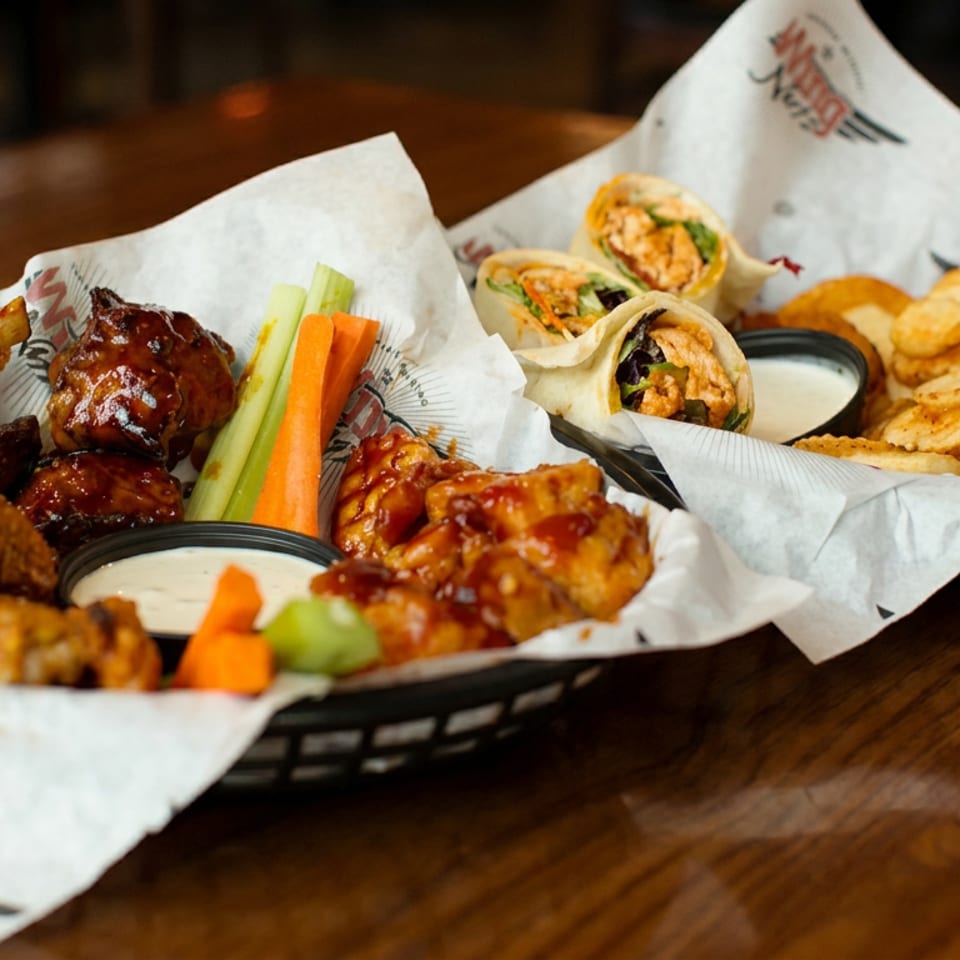 Two baskets hold sauced chicken wings, fried chicken, vegetables, wraps, fries, and dipping sauces on a wooden restaurant table.