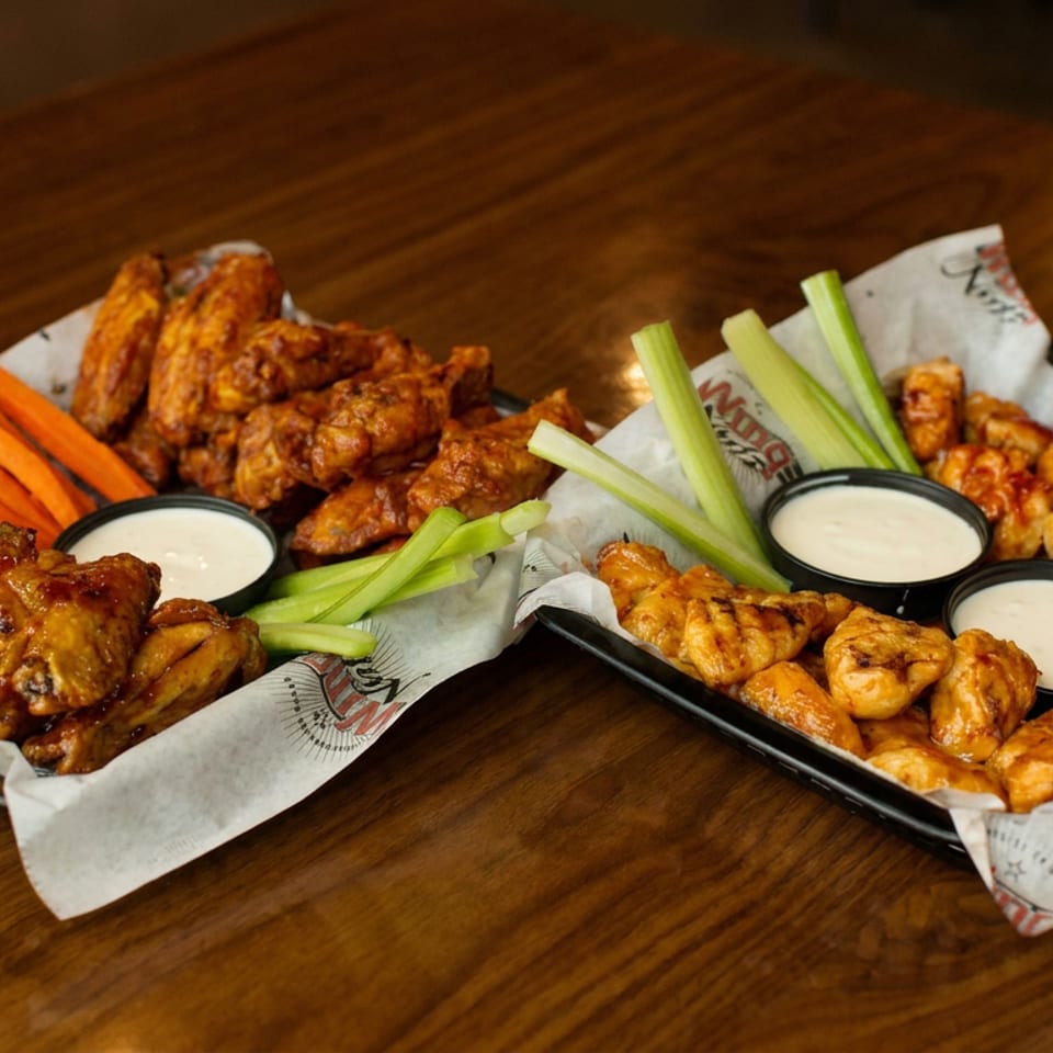 Two trays of saucy chicken wings with carrot and celery sticks, accompanied by cups of creamy dipping sauce on a wooden table.
