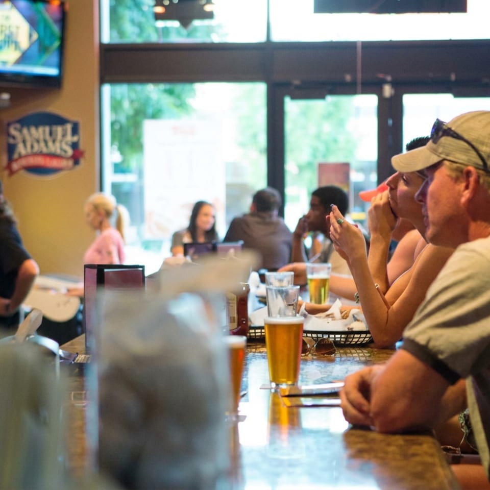 Patrons enjoy drinks and conversation at a lively bar counter beneath a Samuel Adams sign.